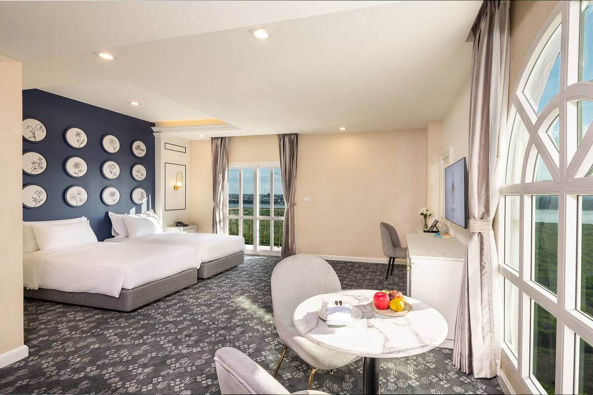 Une chambre d’hôtel avec deux lits, une table à manger avec des fruits, une télévision et une grande fenêtre avec des rideaux.