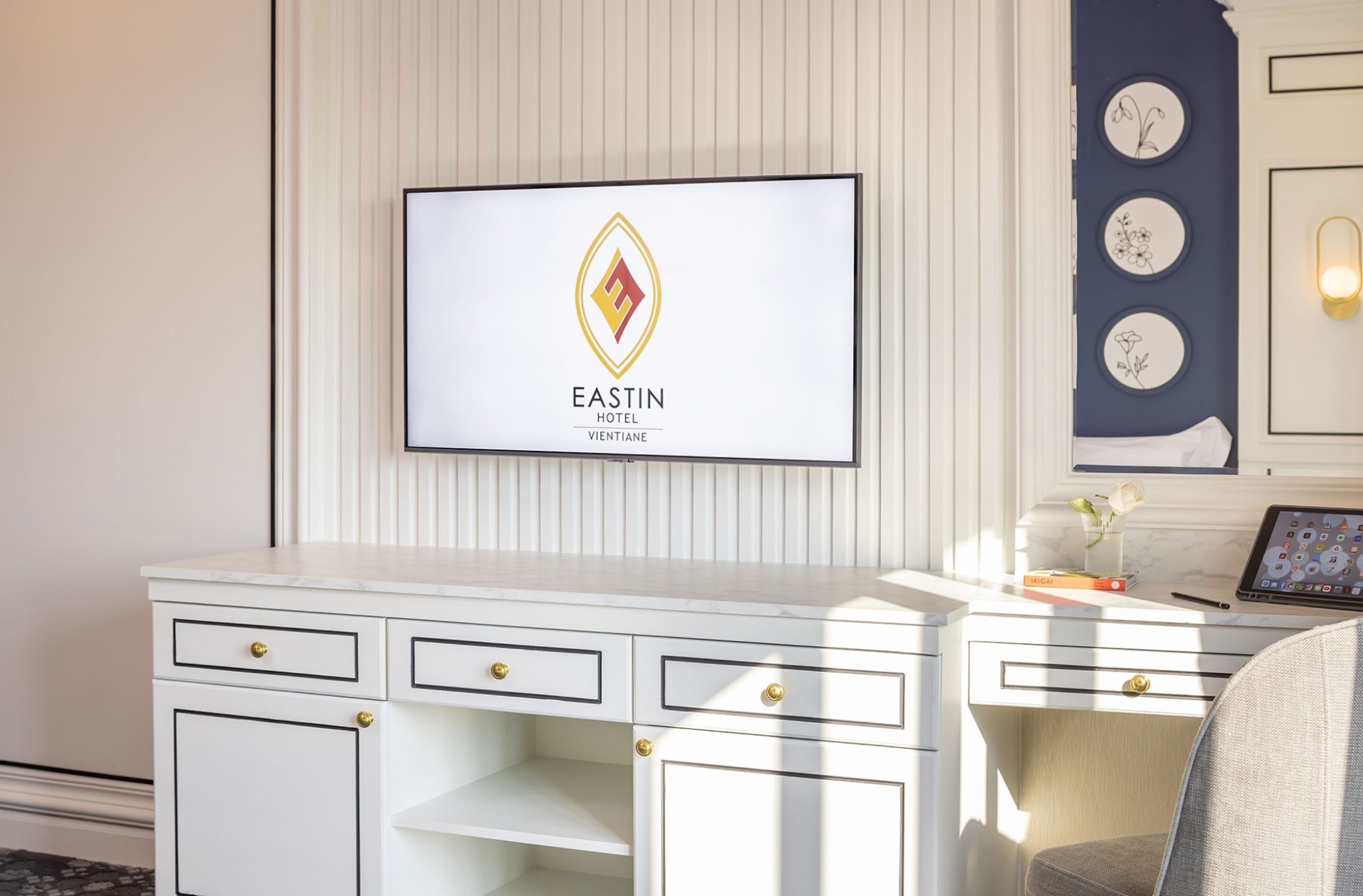 A hotel room with a white desk, a television displaying the Eastin Hotel logo, and a wall with framed artwork.