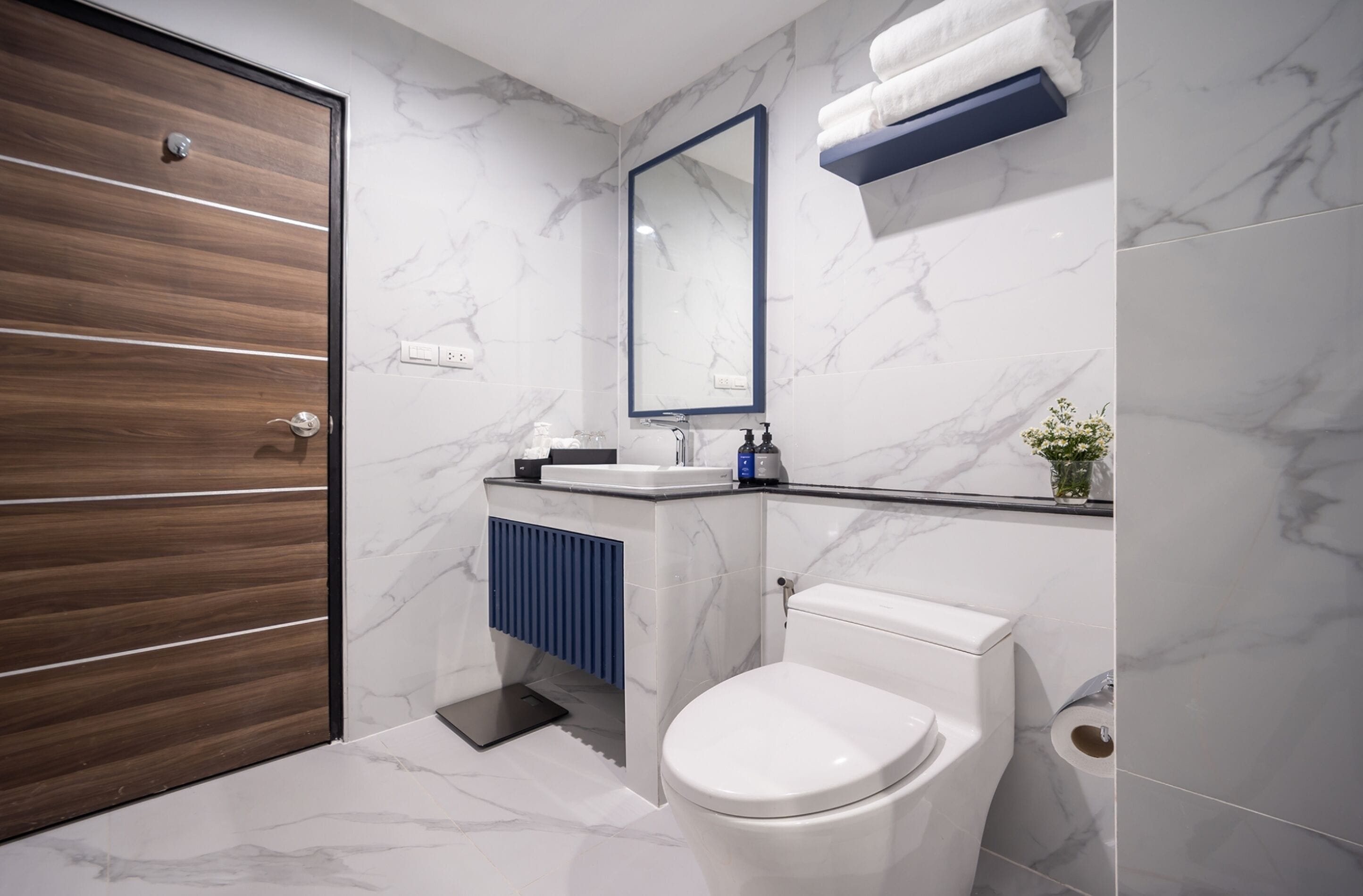 A modern bathroom with a wooden door, white marble walls, a white toilet, a blue vanity, and a mirror above the sink.