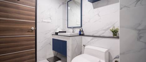 Un baño moderno con puerta de madera, paredes de mármol blanco, inodoro blanco, tocador azul y espejo sobre el lavabo.