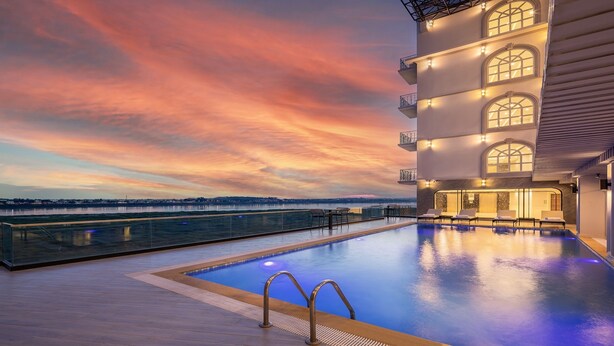 A rooftop infinity pool with a view of the city skyline at sunset, surrounded by modern architecture and outdoor seating.