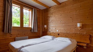 A wooden cabin room with two beds, a window with curtains, and a wall-mounted light fixture.