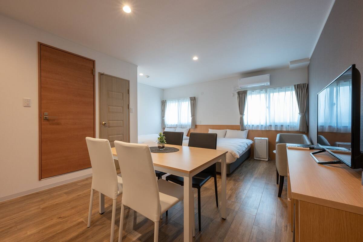 A modern hotel room with a wooden door, a bed with a headboard, a desk with a computer, and a dining area with chairs and a table.