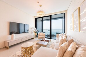 A modern living room with a large TV, comfortable seating, a decorative chandelier, and a view of a cityscape through large windows.