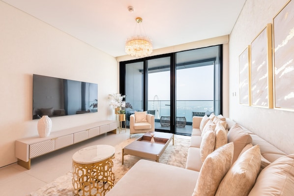 Ein modernes Wohnzimmer mit großem Fernseher, bequemen Sitzgelegenheiten, einem dekorativen Kronleuchter und Blick auf eine Stadtlandschaft durch große Fenster.