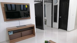 A modern living room with a flat-screen TV mounted on the wall, a wooden entertainment unit, a small coffee table with a plant, and a door leading to another room.