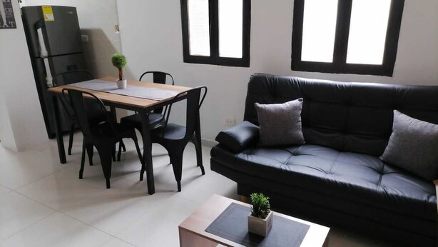 A living room with a black leather sofa, a wooden dining table with chairs, a small potted plant, and a window with blinds.