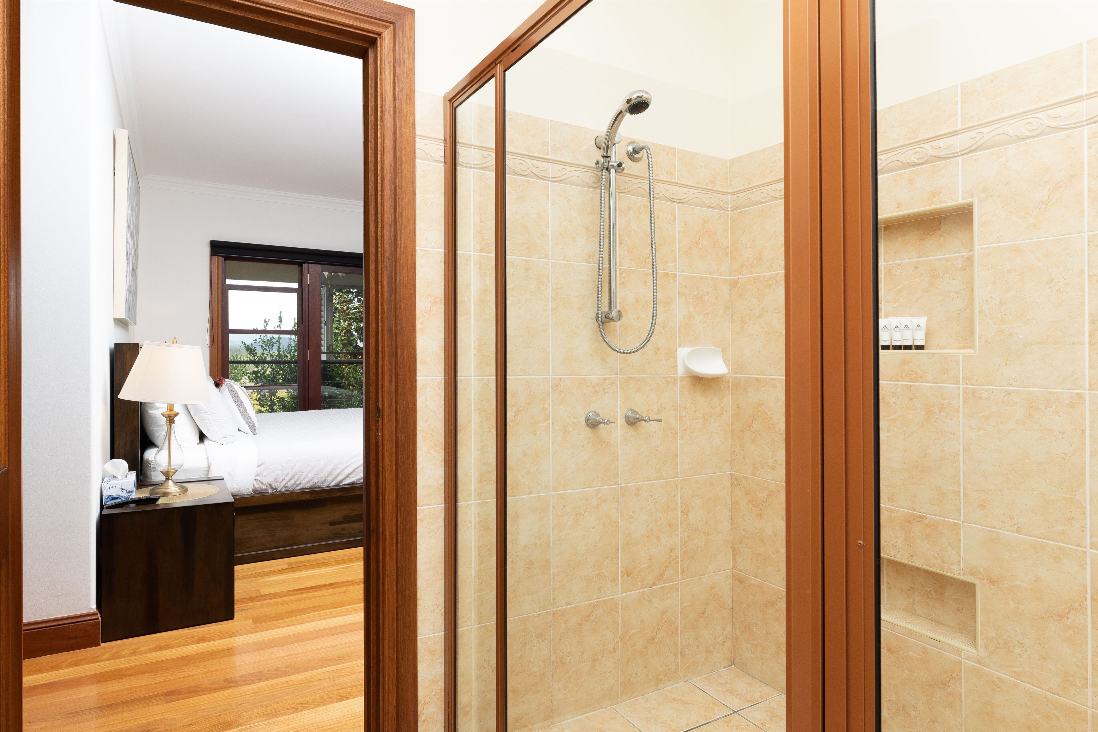 A hotel room with a wooden floor, a bed with white linens, a nightstand with a lamp, and a shower with beige tiles.