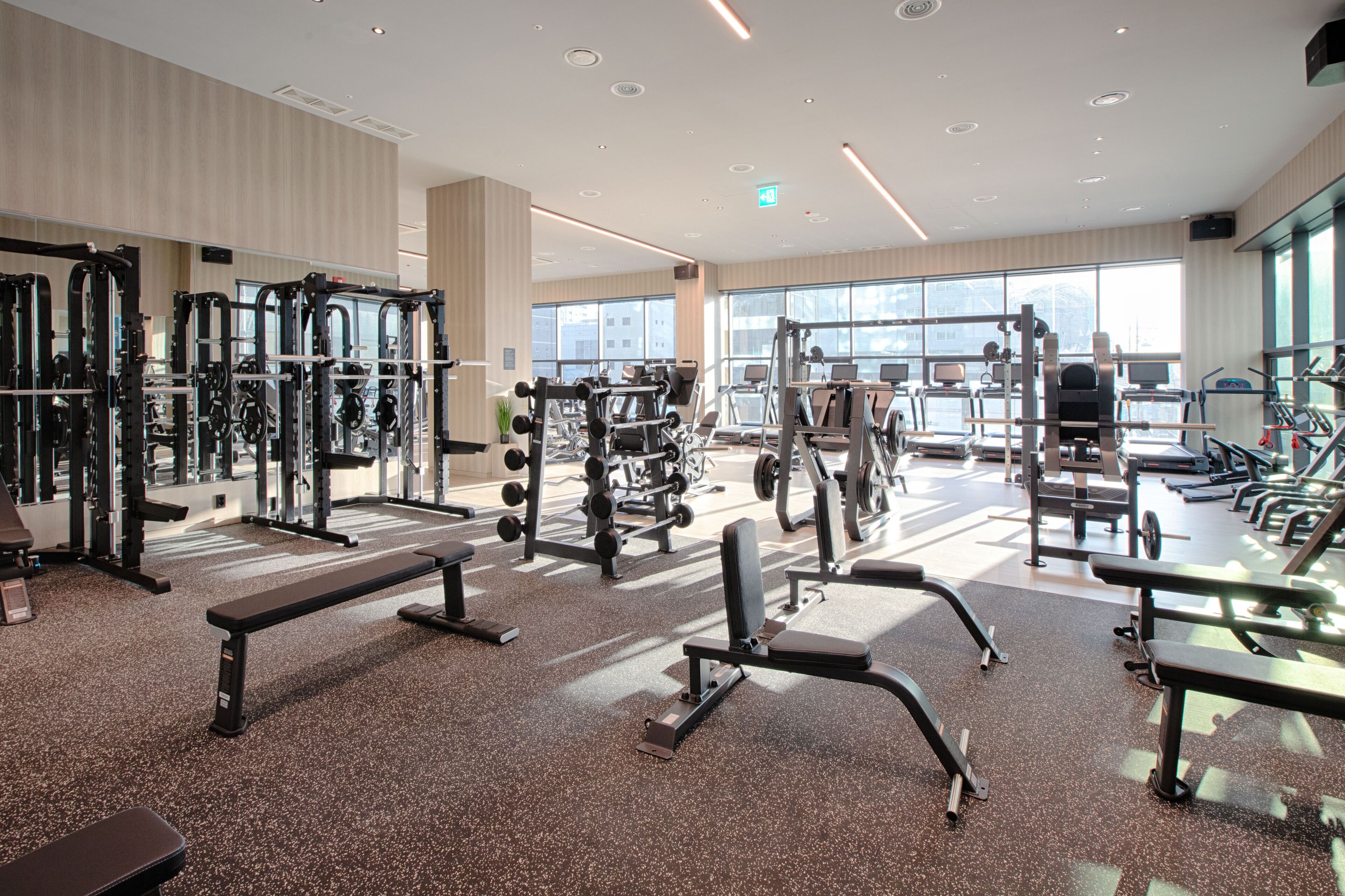 A spacious gym with various exercise equipment, benches, and large windows providing natural light.