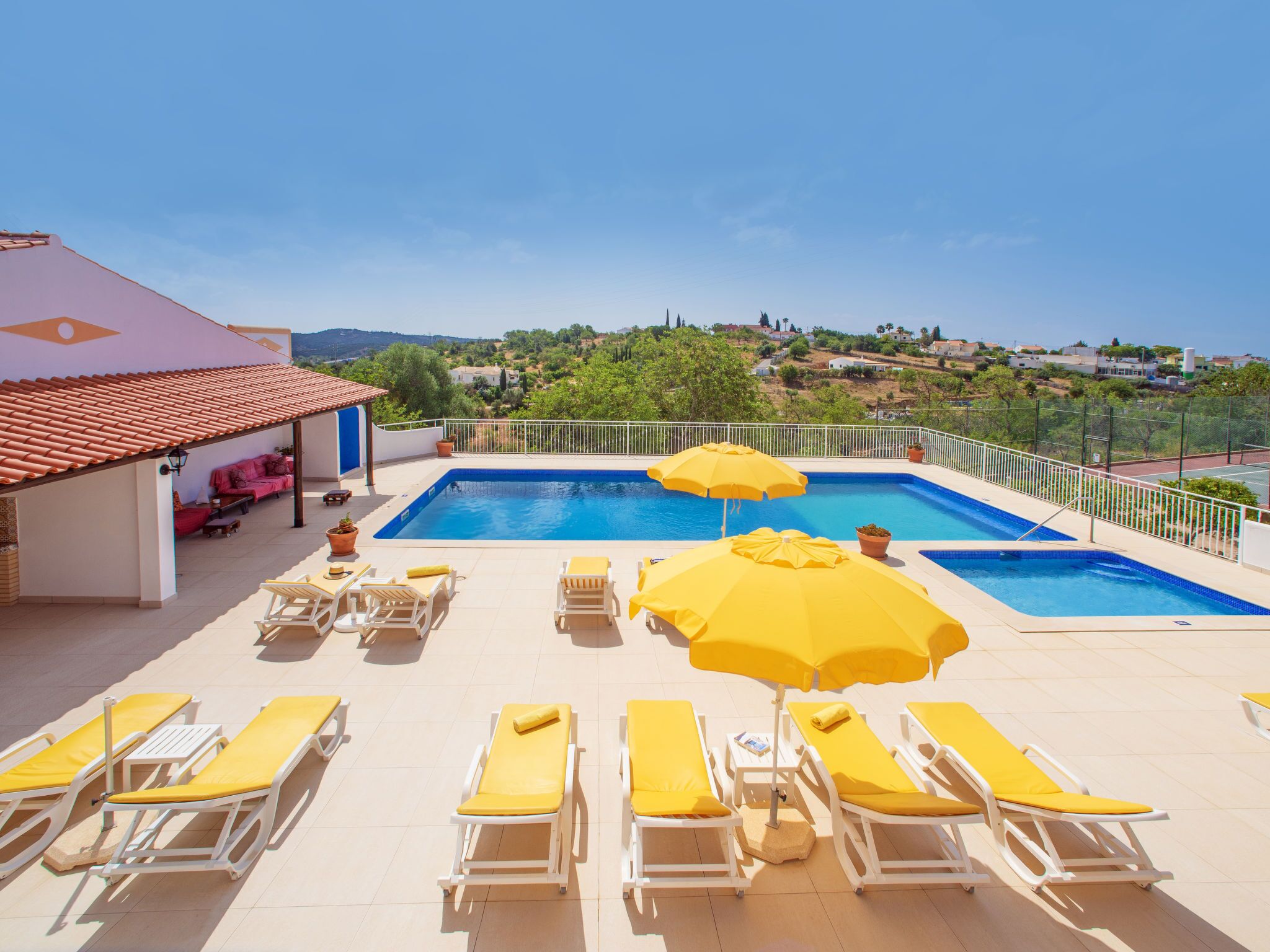 A swimming pool with yellow sun loungers and umbrellas, surrounded by a white fence and overlooking a scenic landscape.