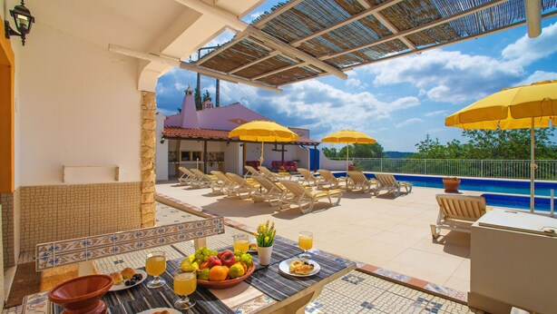 Un espacio al aire libre iluminado por el sol con sillones de descanso y sombrillas amarillas, una piscina y un patio con baldosas con una mesa preparada para el desayuno.