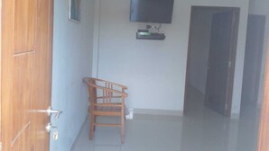 A hallway with a wooden door, a small wooden chair, a flat-screen TV mounted on the wall, and a closed door at the end. - LEMBUNAI HOME STAY (Labuan Bajo)