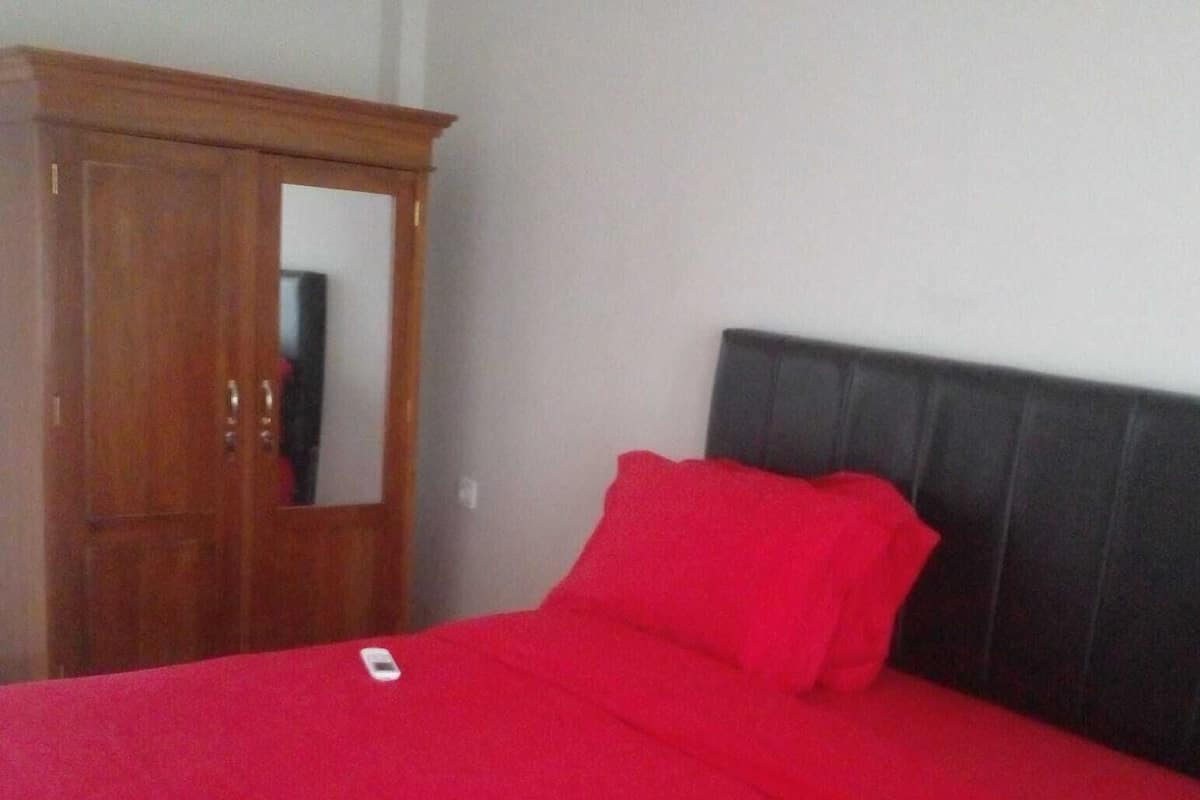 A bed with a red cover and a black headboard, a wooden wardrobe, and a mirror on the wardrobe door.