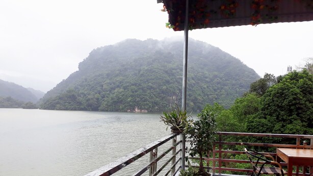 A balcony with a view of a lake and hills. There are potted plants and a table with chairs.