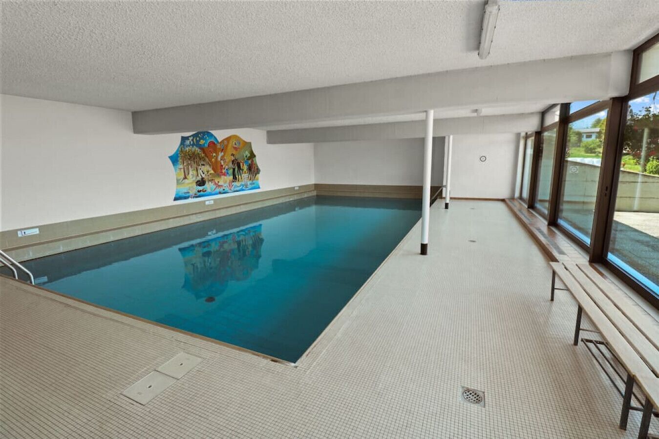 An indoor swimming pool with clear blue water, a tiled floor, and a mural on the wall.