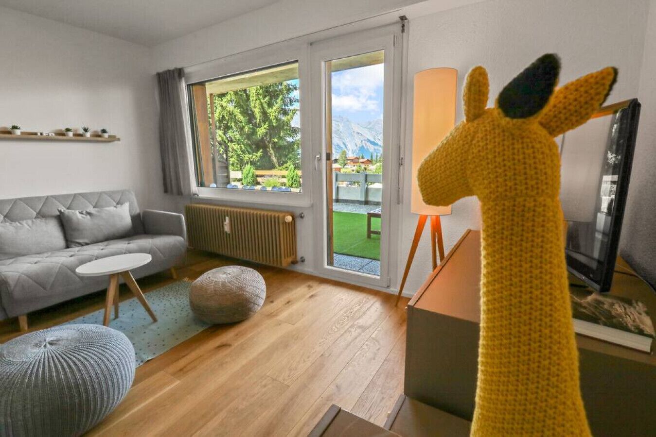 A modern living room with a grey sofa, a round coffee table, a grey pouf, and a large window with a view of greenery and mountains.