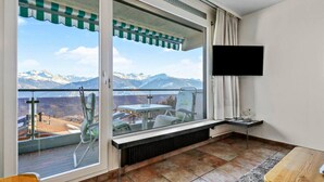 Un balcón con vistas a montañas y un lago, una mesa, sillas y un televisor montado en la pared.