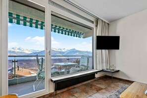 A balcony with a view of mountains and a lake, a table, chairs, and a TV mounted on the wall. - Marigny 65 - Beautiful view, ideal for skiing, quiet location (Crans-Montana)