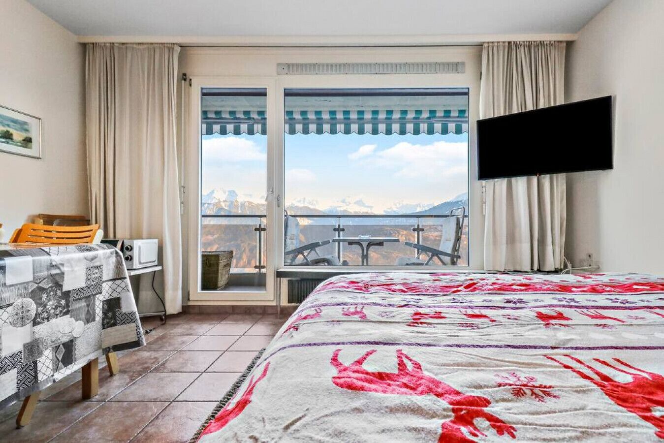 Camera d'albergo con un letto, una scrivania con sedia, una televisione e vista sulle montagne attraverso un'ampia finestra.