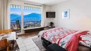A hotel room with a bed, a desk, a chair, a TV, and a large window with a mountain view.