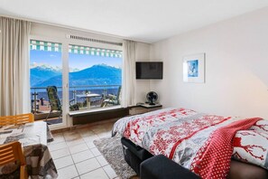 Habitación de hotel con cama, escritorio, silla, televisión y una ventana grande con vista a la montaña.