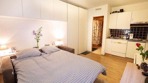 Une chambre à coucher avec un lit, des tables de chevet et une petite table de nuit. On y trouve une petite table sur laquelle est posé un vase de fleurs. La chambre est dotée d’une tête de lit blanche et d’une fenêtre avec des rideaux.