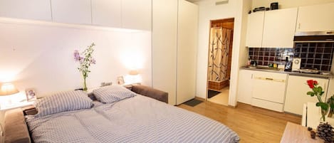 Un dormitorio con una cama, mesitas de noche y una mesita auxiliar. Hay una pequeña mesa con un florero. La habitación tiene un cabecero blanco y una ventana con cortinas.
