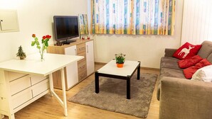 A compact living space with a white desk, a television on a wooden cabinet, a grey sofa with red cushions, and a colorful curtain. - Marigny 45 - Hiking, mountain walks, skiing, quiet location (Crans-Montana)
