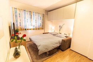 Une chambre à coucher avec un lit, un bureau, une télévision et une fenêtre avec des rideaux.