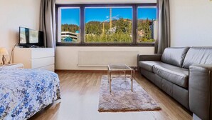 Una habitación de hotel moderna con una cama, un sofá, un televisor y un ventanal que ofrece vistas a un bosque y un edificio.