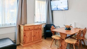 Huoneessa on puinen ruokapöytä, tuoleja, musta toimistotuoli, puinen kaappi ja seinälle asennettu televisio.
