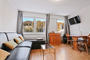 A modern living room with a black leather sofa, a wooden dining table with chairs, a glass coffee table, and a TV mounted on the wall.
