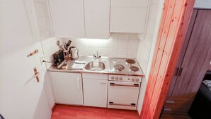 Pieni, käytännöllinen keittiö, jossa on valkoiset kaapit, pesuallas, liesi ja mikroaaltouuni. Oikealla puolella on puinen ovi.
