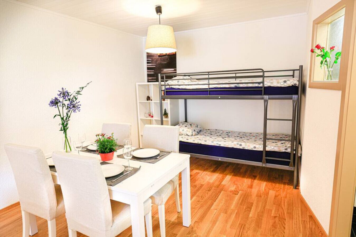 Une pièce de taille modeste, comprenant un lit superposé, une table à manger avec des chaises, une petite plante en pot et un luminaire suspendu.