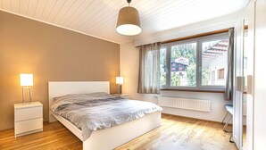 A bedroom with a bed, bedside tables, a window with curtains, a radiator, and a wooden floor. - Clavan 24 - Hiking, mountain walking, quiet area, skiing (Crans-Montana)