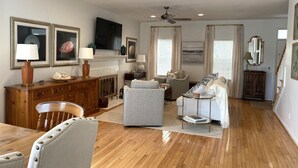 A spacious living room with a wooden floor, a fireplace, a ceiling fan, and a dining area with a wooden table and chairs.