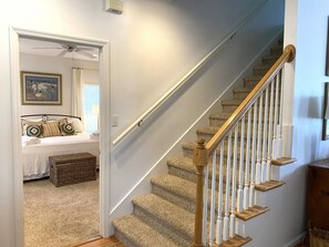 A staircase with carpeted steps and wooden handrails, leading to a bedroom with a bed, pillows, and a framed picture on the wall.