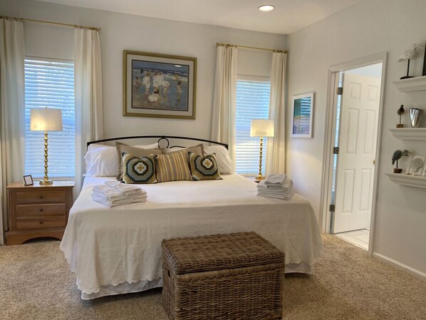 A bedroom with a large bed, two bedside tables, a wicker ottoman, and a painting on the wall.