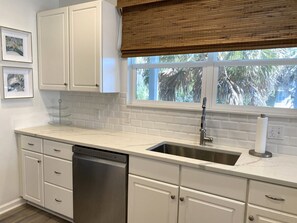 A kitchen with white cabinets, a stainless steel sink, a dishwasher, and a window with a view of palm trees.