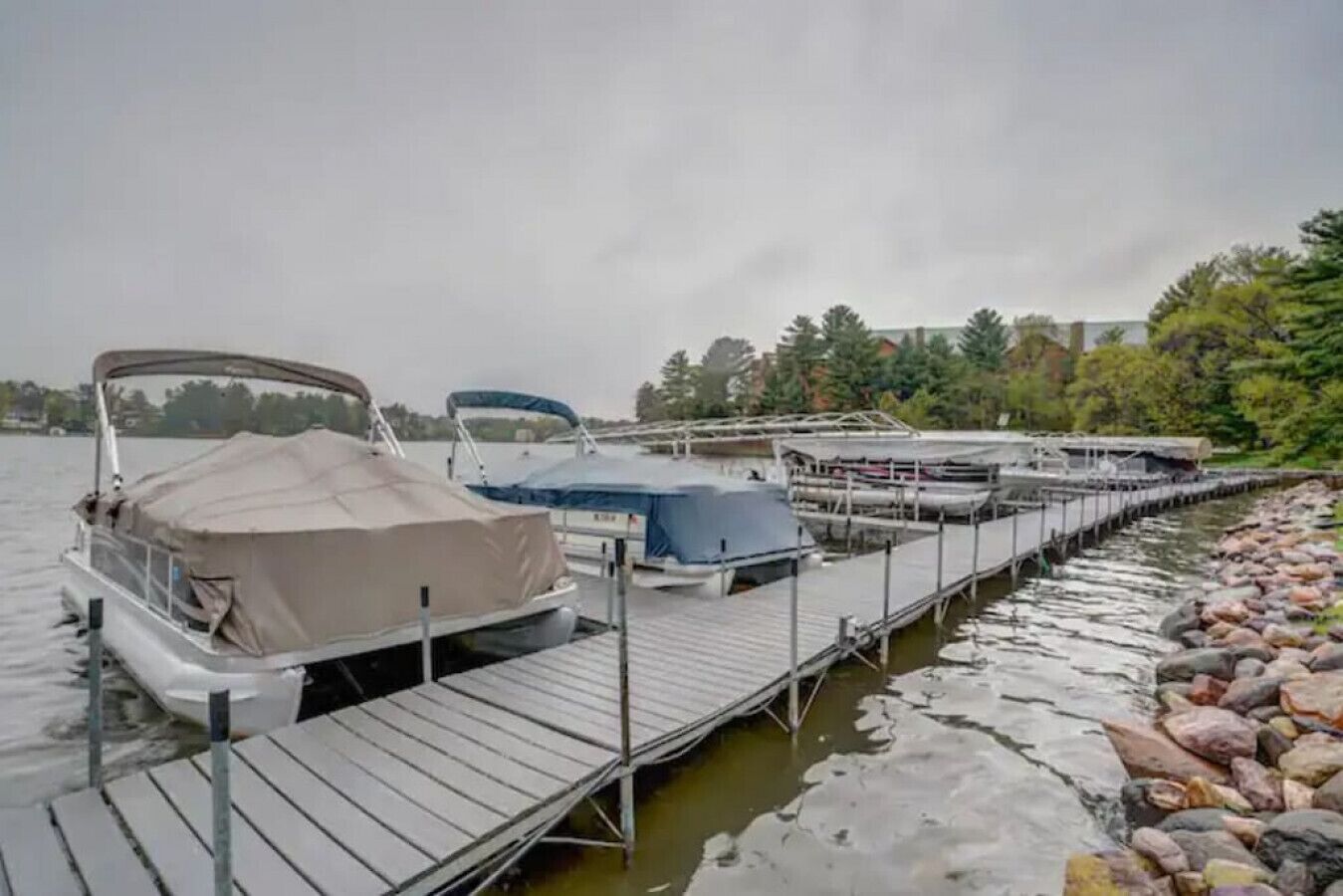 A dock with several boats covered with canopies, a wooden pier, and a rocky shoreline with trees in the background.