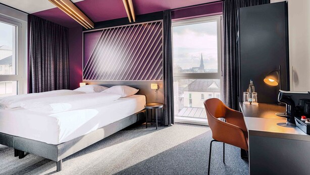A modern hotel room with a large bed, a desk with a chair, a large window with a city view, and a unique ceiling design. - B&B Hotel Bochum-City (Bochum)