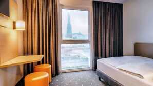 A hotel room with a bed, a desk, two orange ottomans, a large window with a view of a tower, and a wall-mounted TV. - B&B Hotel Bochum-City (Bochum)
