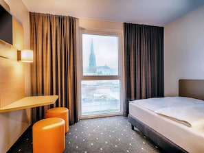 A hotel room with a bed, a desk, two orange ottomans, a large window with a view of a tower, and a wall-mounted TV. - B&B Hotel Bochum-City (Bochum)
