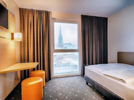 A hotel room with a bed, a desk, two orange ottomans, a large window with a view of a tower, and a wall-mounted TV.