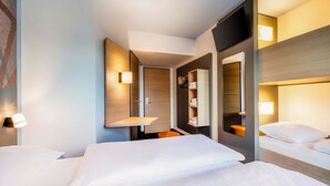 A hotel room with a large bed, a smaller bed, a built-in shelf with towels, a mirror, and a wall-mounted TV. - B&B Hotel Bochum-City (Bochum)