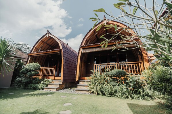 A rustic cabin with a steeply pitched roof, wooden balconies, and a well-maintained garden with various shrubs and a stone pathway. - Gama's Villa (Tabanan)