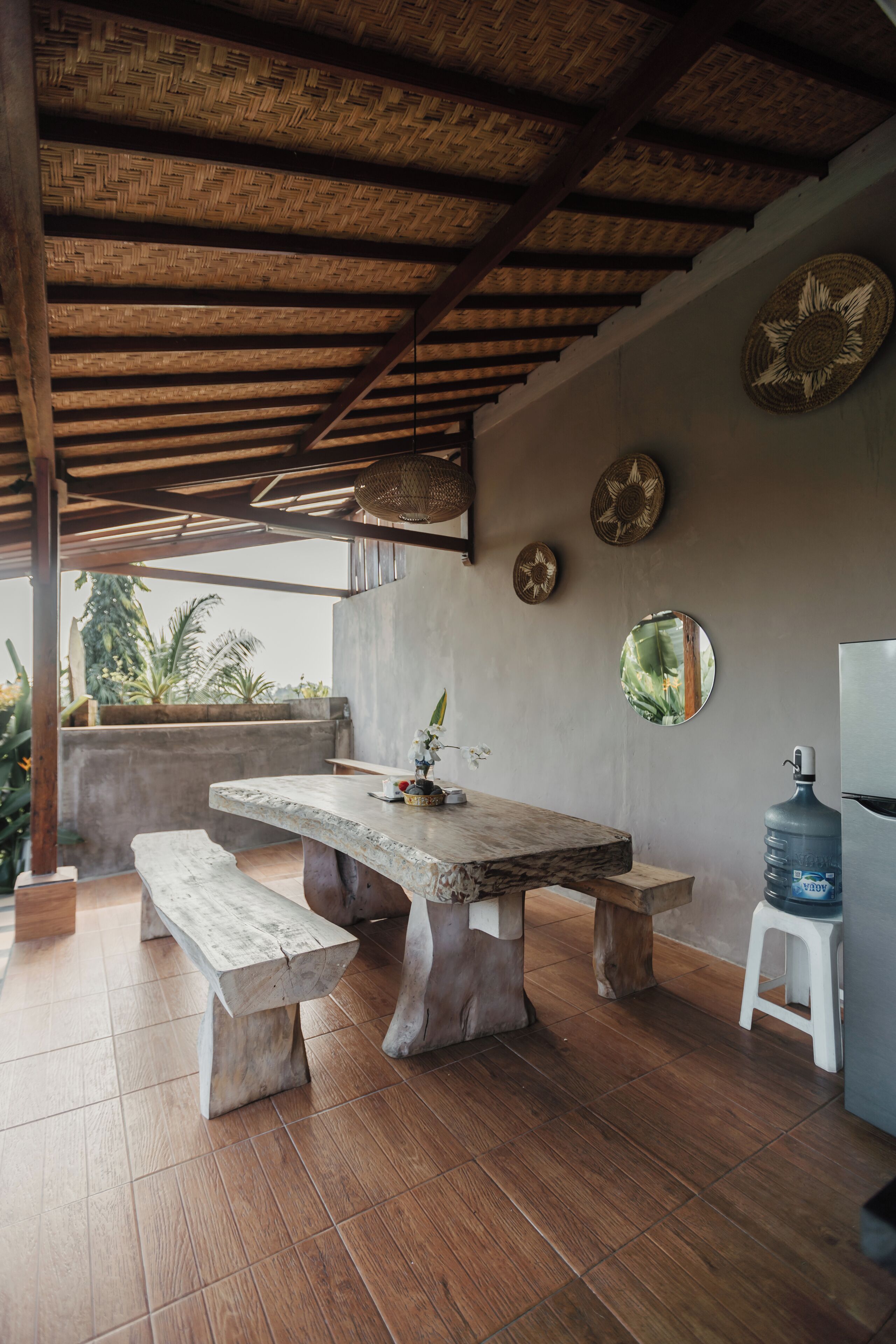 A rustic outdoor dining area with a wooden table and benches, surrounded by a thatched roof and a concrete wall.