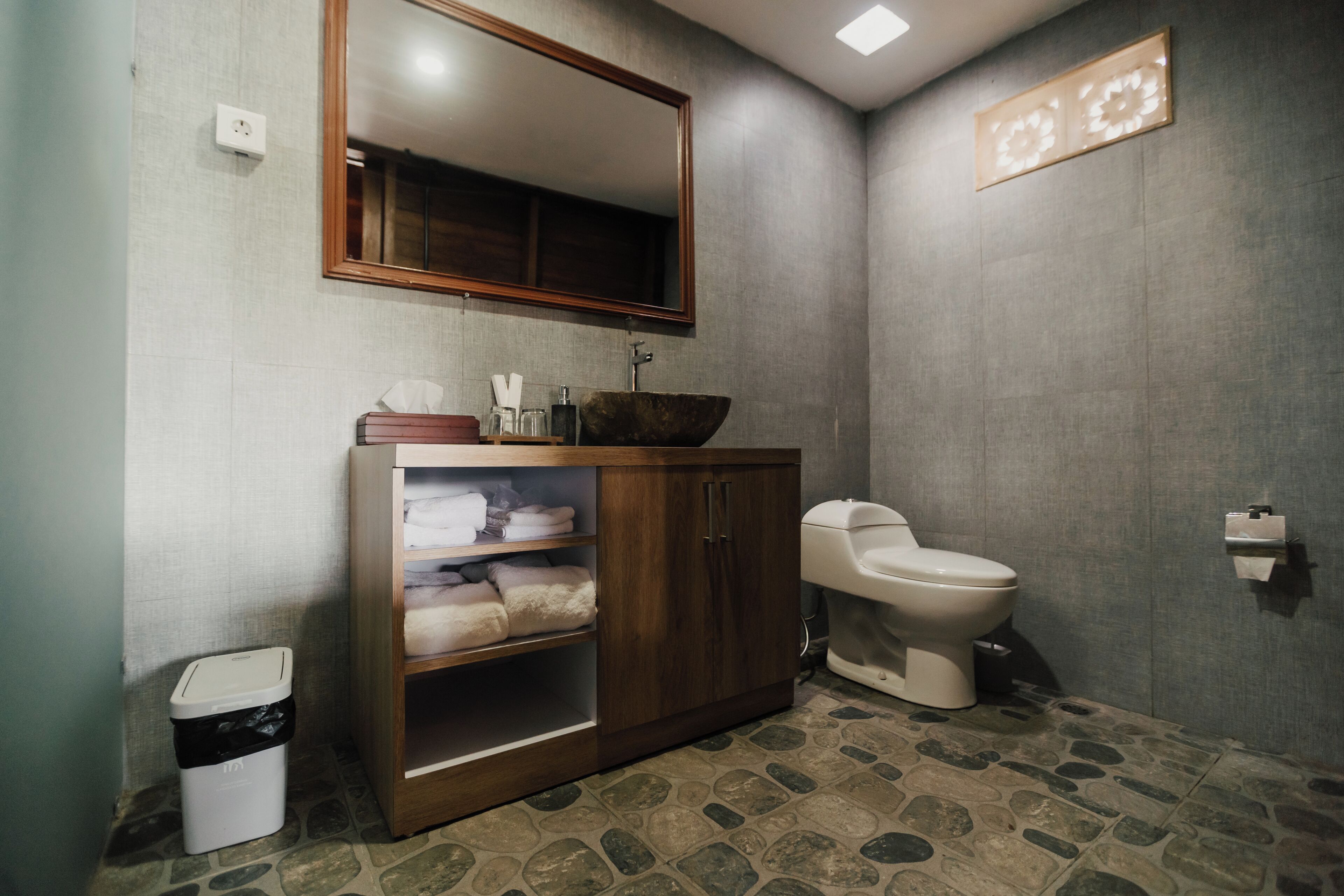 A bathroom with a wooden vanity, a white toilet, a large mirror, and a stone-patterned floor.