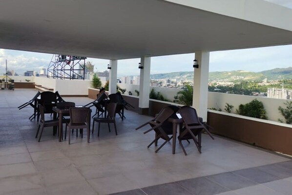 Un espacio al aire libre techado con sillas y mesas, con vista a un paisaje urbano de edificios y zonas verdes.