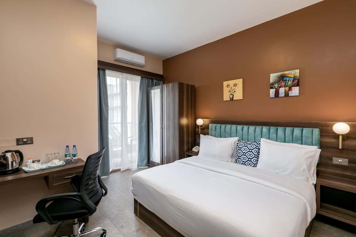A hotel room with a large bed, a desk with a chair, a window with curtains, and two wall-mounted artworks.